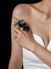A person in a white, strapless top showcases an intricate black and silver Devil's Eye Flower Ring by CAMELLIA, featuring a green stone. Handcrafted in New York, its adjustable size ensures a perfect fit for those who appreciate unique elegance.